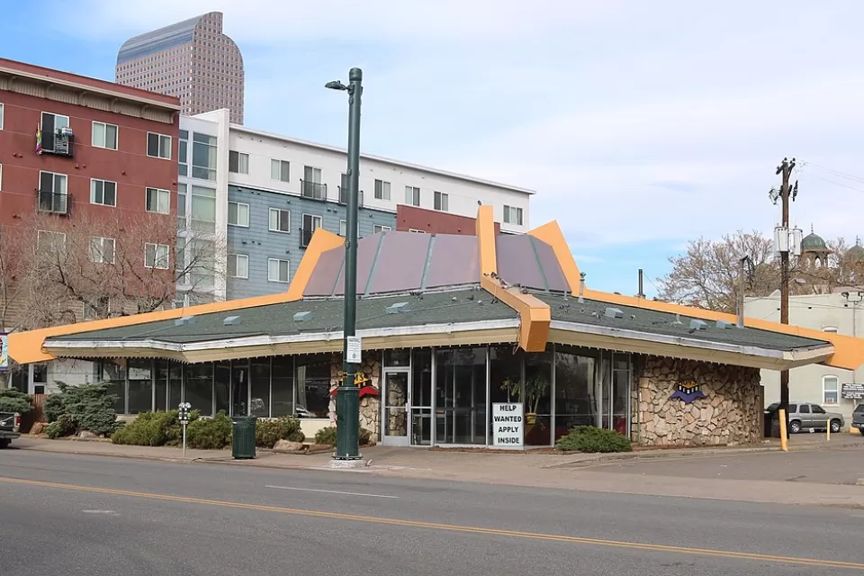 The sloped roof of a googie style building is prominently featured in this image of a restaurant in Denver. Tall apartment buildings are seen in the background.
